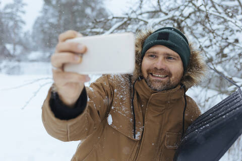Portrait of smiling man with snow shovel taking selfie with cell phone stock photo