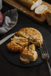 Spanisches Kartoffelomelett, serviert auf einer Brotscheibe und einer alten Silbergabel, aufgenommen aus einem hohen Winkel, mit Brot auf einem Holzbrett im Hintergrund. - CAVF76161