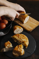 Spanish potato omelette sliced and served with bread. A woman is cutting some bread slices in the background. - CAVF76162