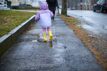 Ein kleines Mädchen, das an einem regnerischen Tag in Pfützen planscht. - CAVF76238