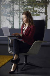 Geschäftsfrau sitzt im Sessel im Büro und benutzt einen Laptop - RBF07082