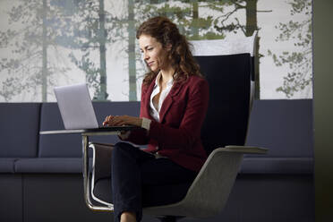 Geschäftsfrau sitzt im Sessel im Büro und benutzt einen Laptop - RBF07083