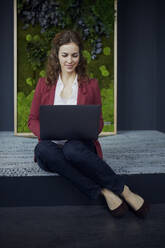 Smiling businesswoman sitting on the floor in green office using laptop - RBF07086