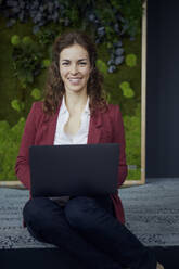 Portrait of smiling businesswoman sitting on the floor in green office using laptop - RBF07087