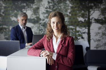 Porrait einer lächelnden Geschäftsfrau im Büro mit einem Geschäftsmann im Hintergrund - RBF07095