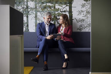 Geschäftsmann und Geschäftsfrau sitzen auf einer Couch im Büro und teilen ein Tablet - RBF07097