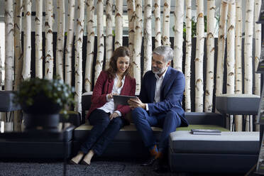 Businessman and businesswoman sitting on couch in office sharing tablet - RBF07106