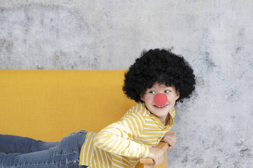 Junge mit roter Clownsnase und schwarzen Haaren auf gelber Couch - HMEF00785