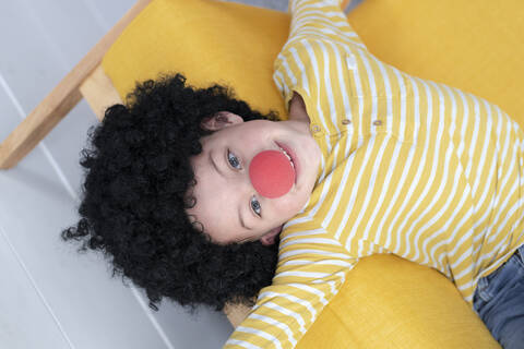 Junge mit roter Clownsnase und schwarzen Haaren auf gelber Couch, lizenzfreies Stockfoto