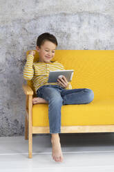 Junge spielt mit Tablet auf gelber Couch - HMEF00788