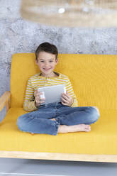 Junge spielt mit Tablet auf gelber Couch - HMEF00792