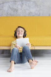 Boy playing with tablet and wearing headphones at a yellow couch - HMEF00796