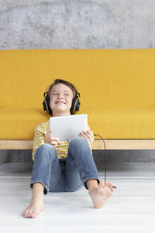 Junge spielt mit Tablet und trägt Kopfhörer auf einer gelben Couch - HMEF00796