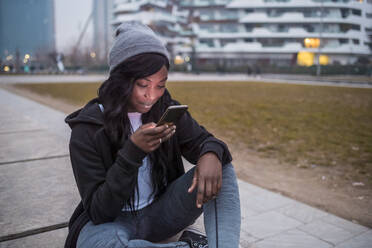 Lächelnde junge Frau, die in der Abenddämmerung in der Stadt ihr Smartphone benutzt - MEUF00040