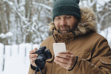 Portrait of bearded man looking with digital camera looking at cell phone in winter - KNTF04482