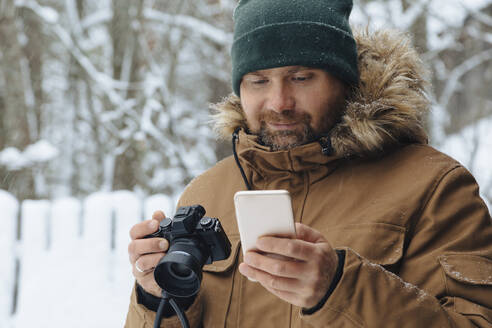 Porträt eines bärtigen Mannes, der mit einer Digitalkamera auf ein Handy im Winter blickt - KNTF04482