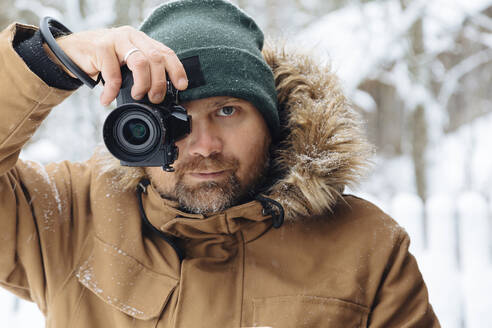 Porträt eines bärtigen Mannes, der den Betrachter mit einer Digitalkamera im Winter fotografiert - KNTF04484