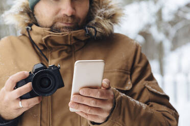 Crop view of man with digital camera looking at cell phone in winter - KNTF04485
