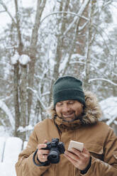 Portrait of relaxed man with digital camera and mobile phone in winter forest - KNTF04489