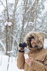 Porträt eines entspannten Mannes mit Digitalkamera und Mobiltelefon im Winterwald - KNTF04491