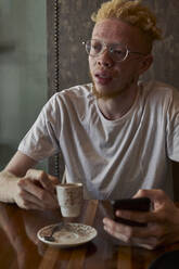 Albino-Mann mit runder Brille, der in einer Cafeteria einen Kaffee trinkt und sein Telefon überprüft - VEGF01652