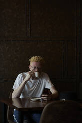 Albino-Mann mit runder Brille, der in einer Cafeteria einen Kaffee trinkt und sein Telefon überprüft - VEGF01653