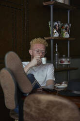 Entspannter Albino-Mann, der einen Kaffee trinkt und die Beine auf den Tisch legt - VEGF01660
