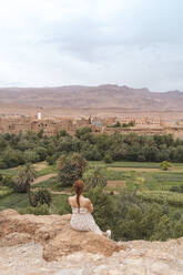 Rückenansicht einer jungen Frau, die auf einem Felsen sitzt und auf die Stadt blickt, Ouarzazate, Marokko - AFVF05569
