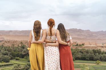 Rückenansicht von drei jungen Frauen, die Arm in Arm stehen und auf die Stadt blicken, Ouarzazate, Marokko - AFVF05570