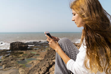 Junge Frau sitzt am Meer und schaut auf ihr Handy, Essaouira, Marokko - AFVF05590