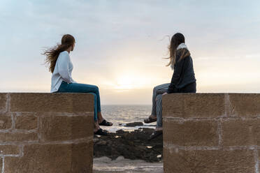 Zwei junge Frauen sitzen auf einer Mauer am Strand und beobachten den Sonnenuntergang, Essaouira, Marokko - AFVF05595