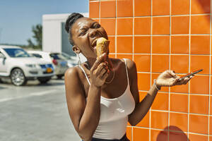 Junge lachende Frau, die ein Eis isst und ihr Handy überprüft - VEGF01670