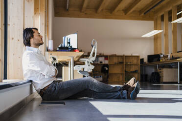 Businessman with closed eyes sitting at the window in open-plan office - DIGF09404