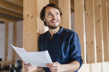 Smiling businessman holding documents in wooden open-plan office - DIGF09443