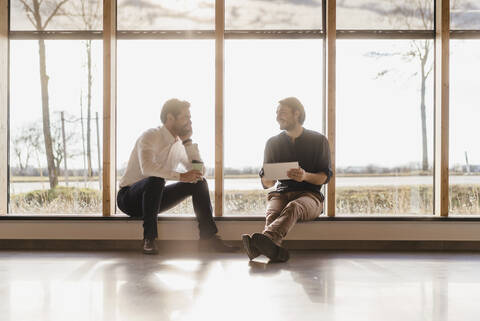 Zwei Geschäftsleute mit Tablet im Gespräch am Fenster in einem Großraumbüro, lizenzfreies Stockfoto