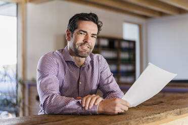 Portrait of smiling businessman with document in wooden open-plan office - DIGF09493