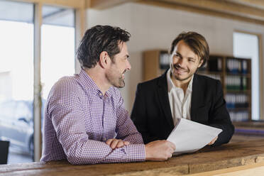 Zwei lächelnde Geschäftsleute mit Dokumenten im Gespräch in einem hölzernen Großraumbüro - DIGF09496
