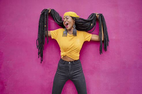 Portrait of woman with long dreadlocks holding her hair in front of a pink wall - VEGF01680