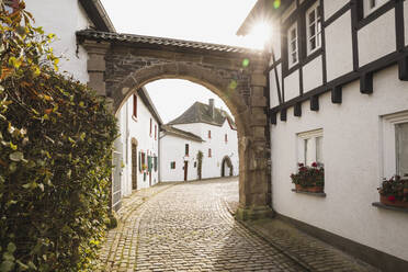 Germany, North Rhine-Westphalia, Reifferscheid, Sun shining over archway in empty cobblestone alley - GWF06517