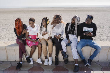 Happy teenage girlfriends and young man sitting on a wall at the beach with smartphones - MPPF00595