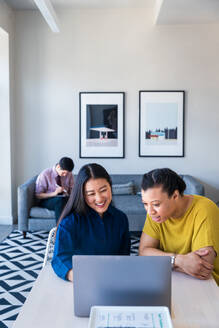 Lächelnde Unternehmerinnen planen eine Strategie am Laptop, während ein männlicher Kollege auf dem Sofa im Büro sitzt - CAVF76475