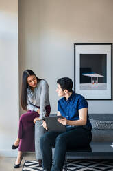 Männliche und weibliche Mitarbeiter planen über einem Grafiktablett auf einem Sofa an der Wand im Büro - CAVF76494