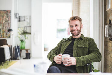 Portrait of smiling male entrepreneur with cup sitting at home office - MASF16853