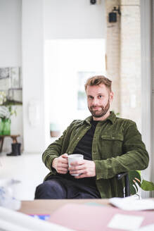 Porträt eines männlichen Unternehmers mit einer Tasse, der im Büro zu Hause sitzt - MASF16855