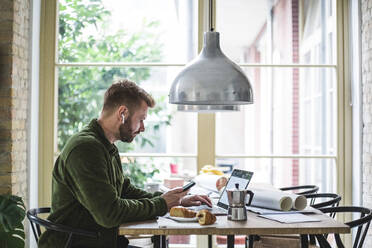 Male entrepreneur using laptop while holding phone at home office - MASF16883