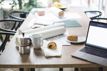 High angle view of teapot with documents rolled up by laptop and croissant on table - MASF16890