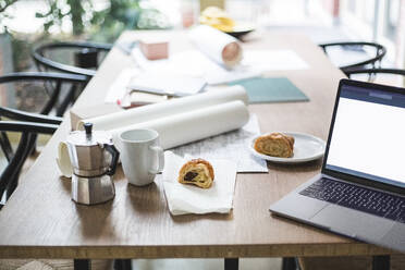 Hochformatige Ansicht einer Teekanne mit aufgerollten Dokumenten neben Laptop und Croissant auf dem Tisch - MASF16890