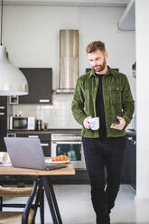 Männlicher Architekt mit Kaffeetasse, der an einem Tisch zu Hause vorbeigeht - MASF16905