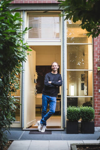 Porträt eines männlichen Architekten, der mit verschränkten Armen vor einem Hauseingang steht, lizenzfreies Stockfoto