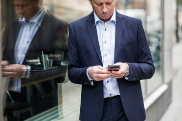 Contemplating mature businessman using smart phone while standing by glass window - MASF16927